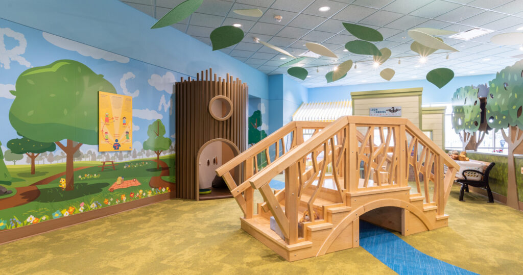 A wooden bridge on a blue and green carpet with fake leaves hanging from the ceiling and a tree trunk structure in the background.